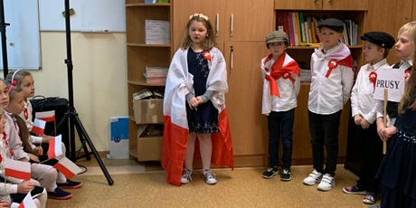 Powiększ grafikę: Dziewczynka okryta flagą polski mówi wiersz. Obok stoją uczniowie którzy są ubrani na galowo.