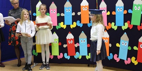 Powiększ grafikę: Pierwszoklasistka mówi wiersz obok  niej stoją dwie uczennice z klasy 5. Na drugim planie widać wychowawcę. 